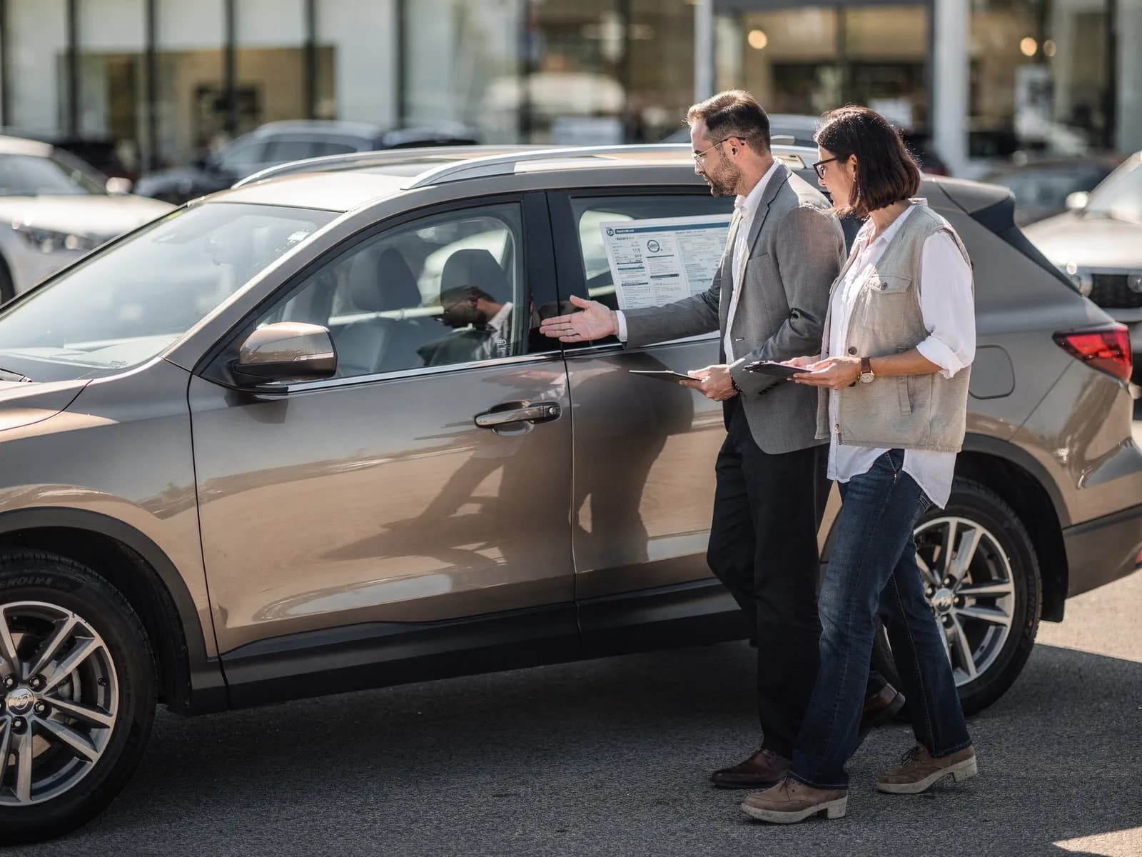Salesperson and customer walking a dealership lot beside a parked SUV, both looking at a printed window sticker