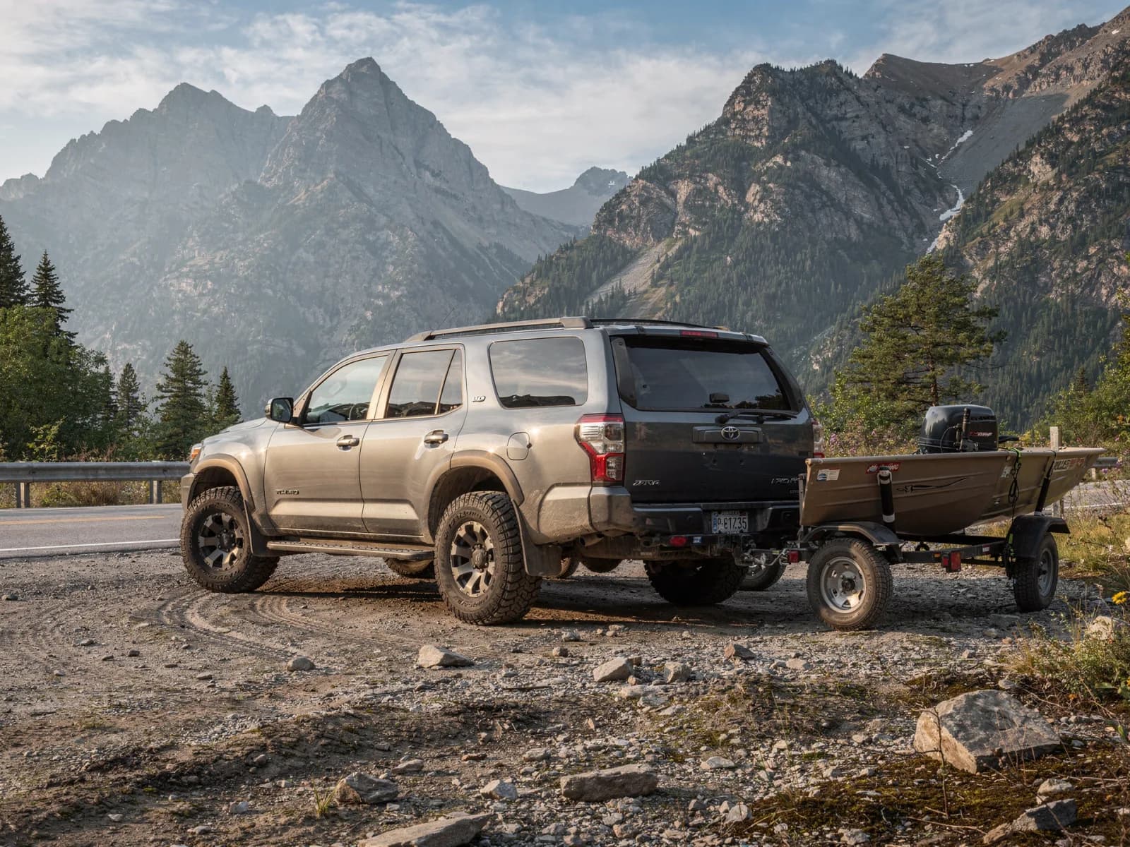 Grey full-size body-on-frame SUV parked on a gravel pull-off with a boat trailer hitched, mountains behind