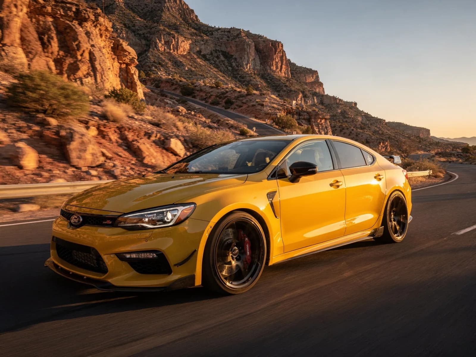 Yellow compact rear-drive sport sedan on a canyon road in low sun with visible brake calipers behind the wheels