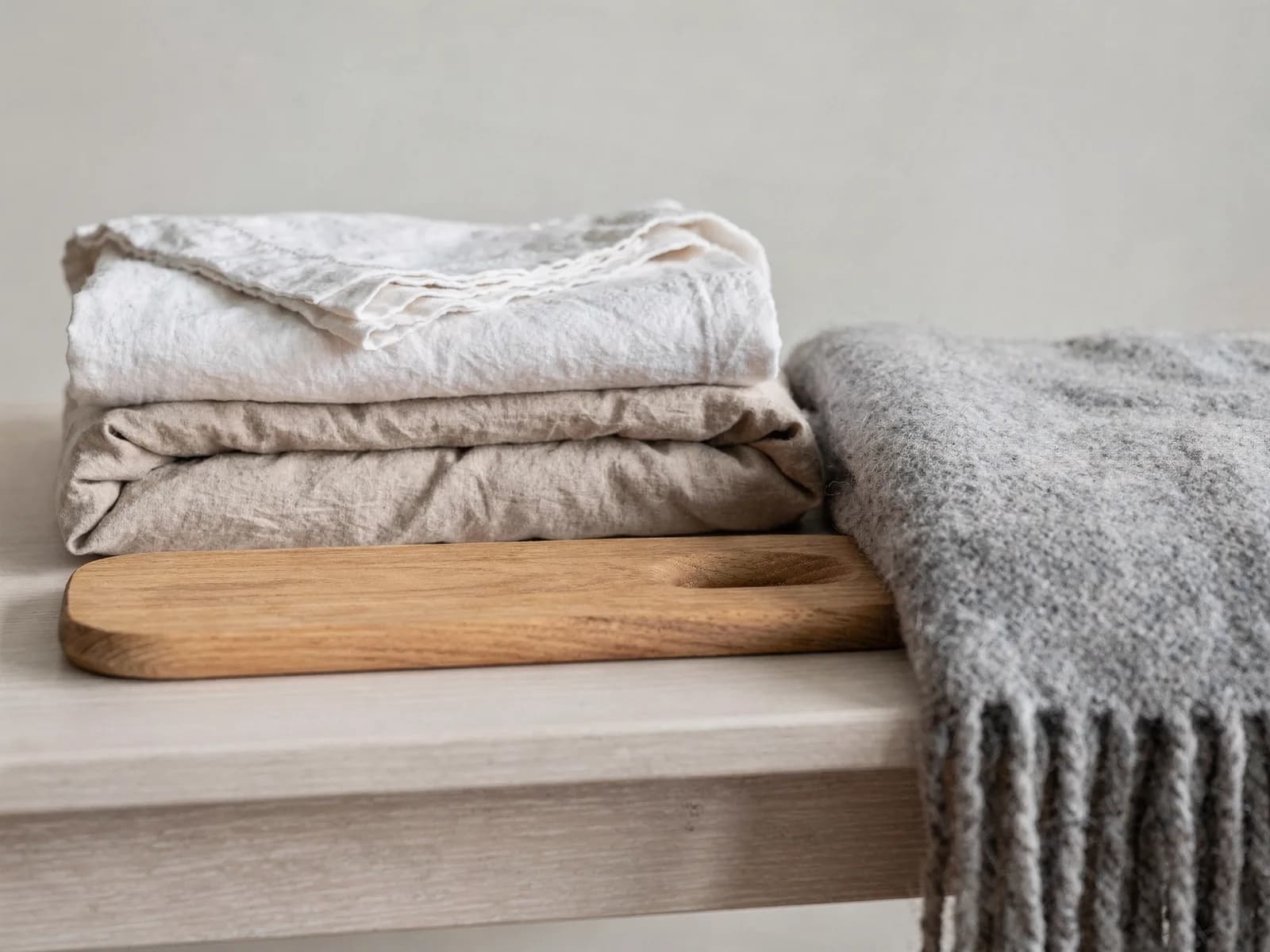 Still life of a folded wool throw, stacked linen bedding and an oak serving board on a pale wooden table