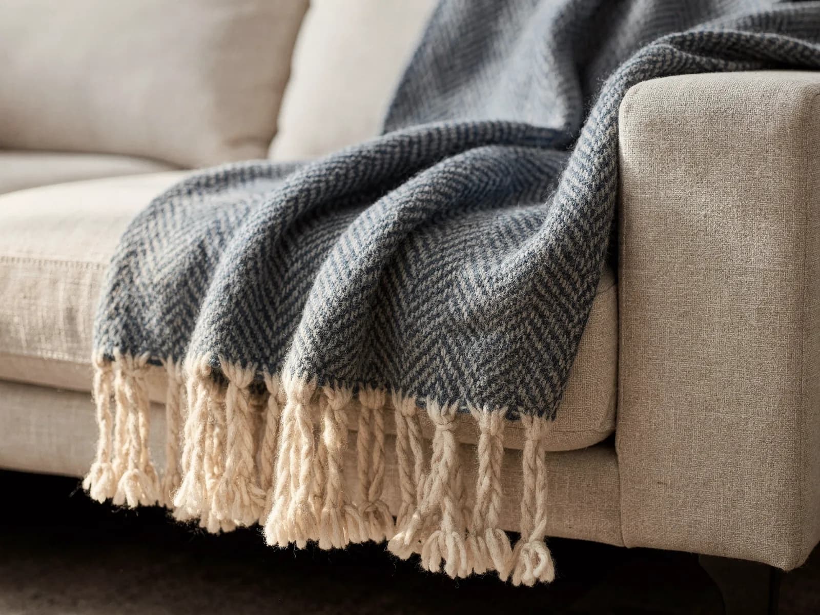 Heavy blue herringbone lambswool throw draped over the arm of a linen sofa with the knotted fringe visible